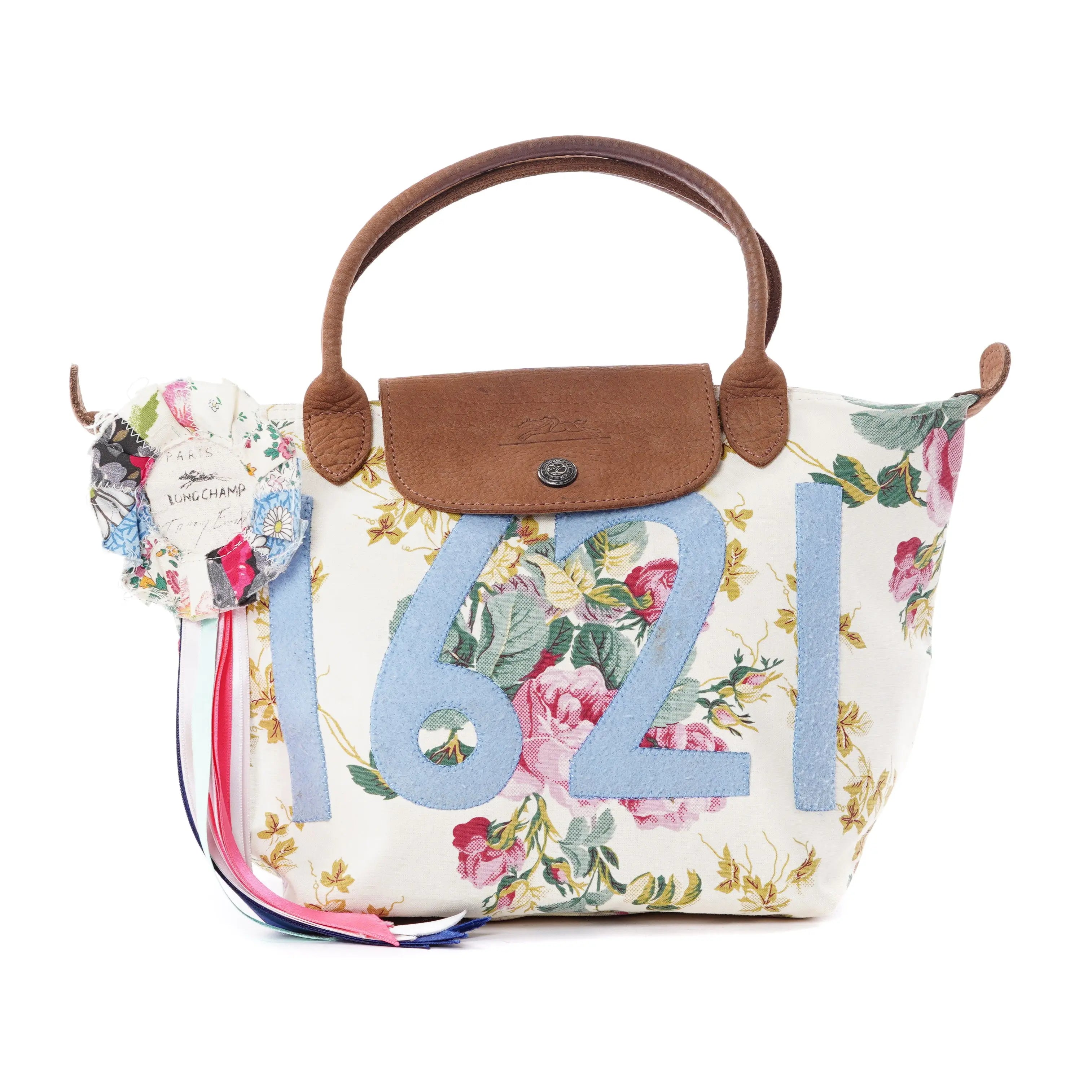 Longchamp Pliage Tracey Emin handbag features a floral design with brown handles and a flap. It showcases large blue 1621 numbers on the front, resembling Tracey Emins work, and includes a decorative round patch with ribbons on one side.